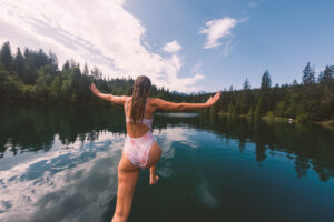 Young woman in swimsuit jumps into calm lake with arms outstretched. Scenic landscape with serene waters. Leisure and nature adventure in summer.